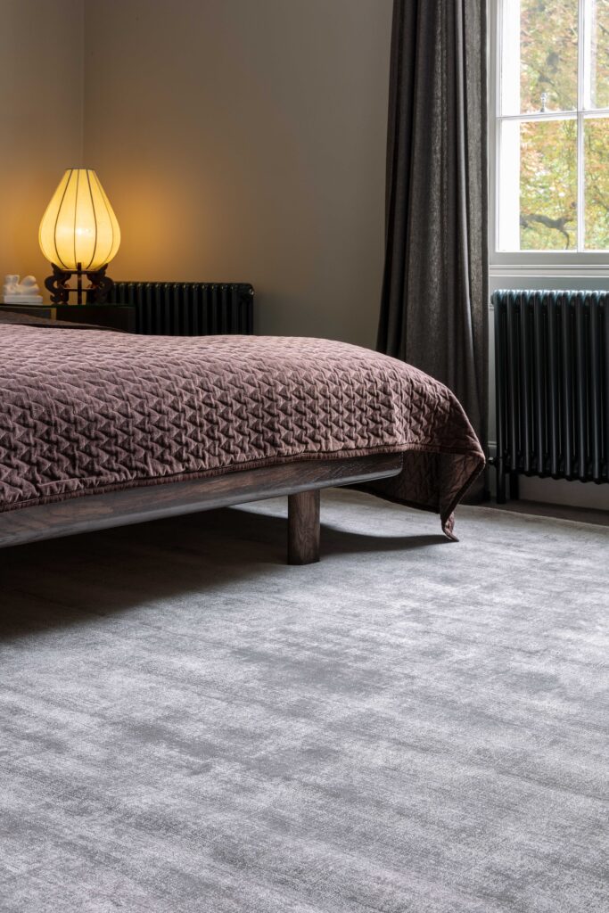 Tappeto Blade Silver di Asiatic Carpets in viscosa, posizionato in una camera da letto elegante con letto in legno e copriletto marrone