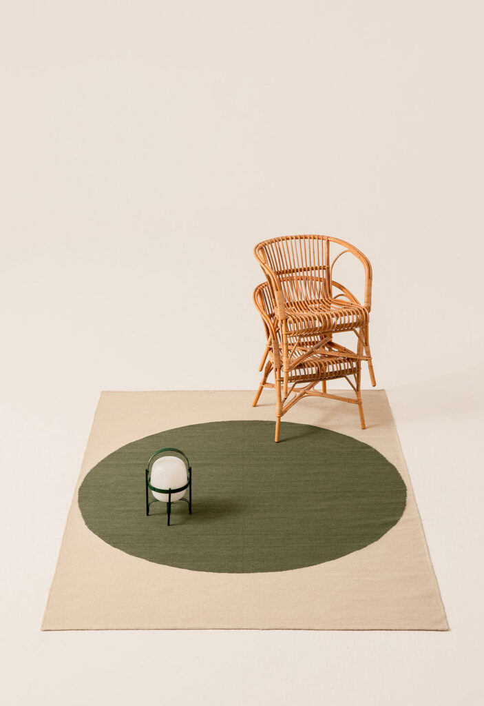 Tappeto Topo verde smeraldo di Nanimarquina con cerchio decentrato, lanterna moderna e sedie impilate in rattan su sfondo chiaro